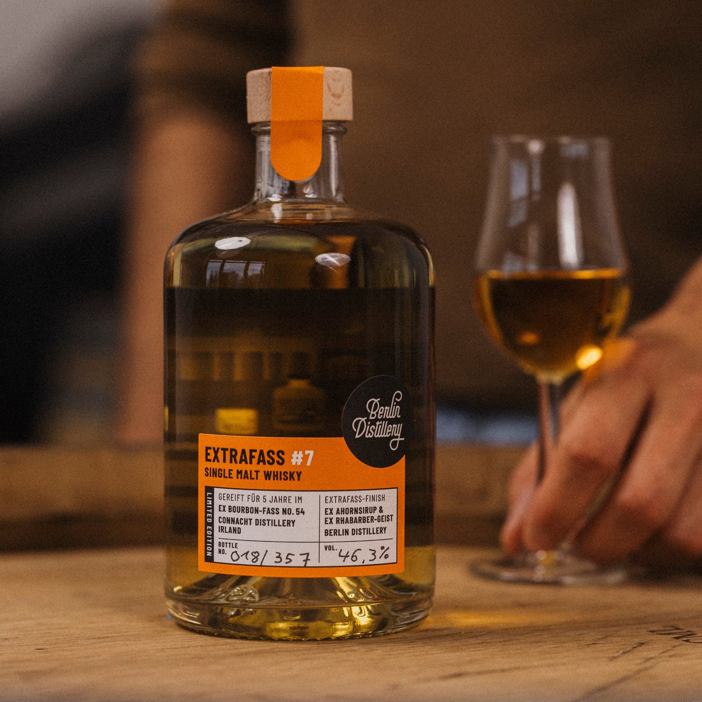 Eine Flasche EXTRAFASS #7 Single Malt Whisky der Berlin Distillery steht auf einem Whiskyfass. Daneben greift eine Hand ein volles Whiskyglas.