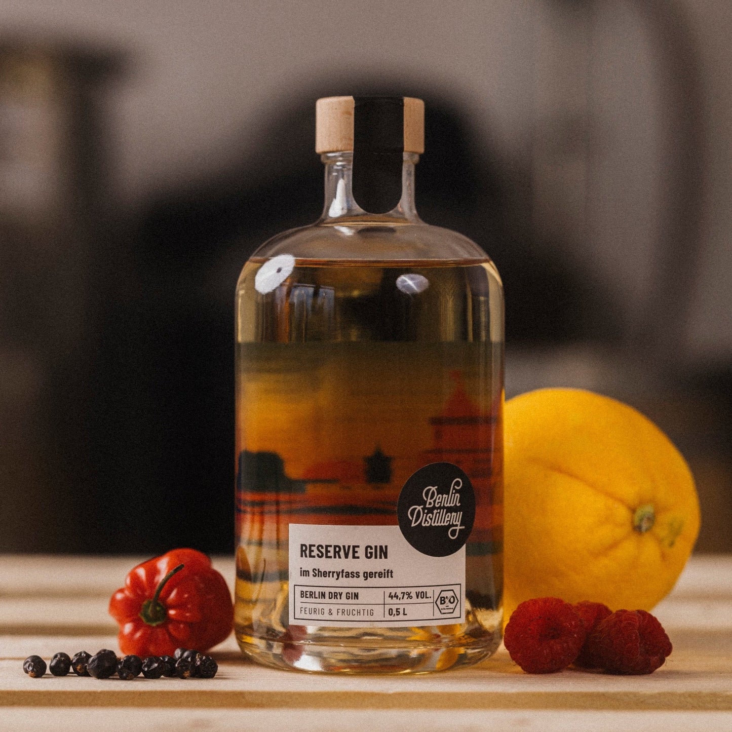 Eine Flasche Berlin Distillery Reserve Gin steht zwischen einer Zitrone, einer Habanero-Chili, Himbeeren und Pfeffer auf einer Holzpalette vor dem Hintergrund der Produktionseinrichtung.