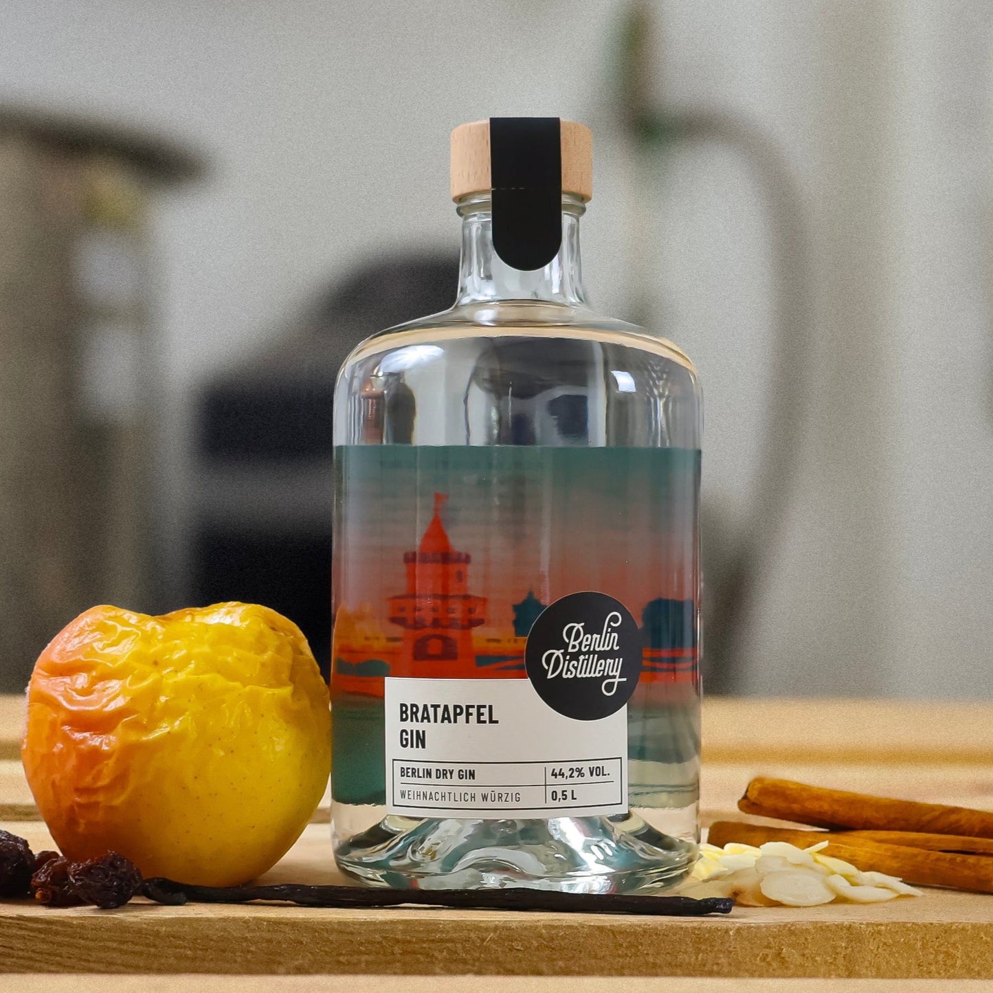 Eine Flasche Berlin Distillery Bratapfel Gin steht auf einer Holzpalette vor der Produktionsanlage. Neben der Glasflasche liegen ein Bratapfel, Rosinen, gehobelte Mandeln, eine Vanilleschote und Zimtstangen.