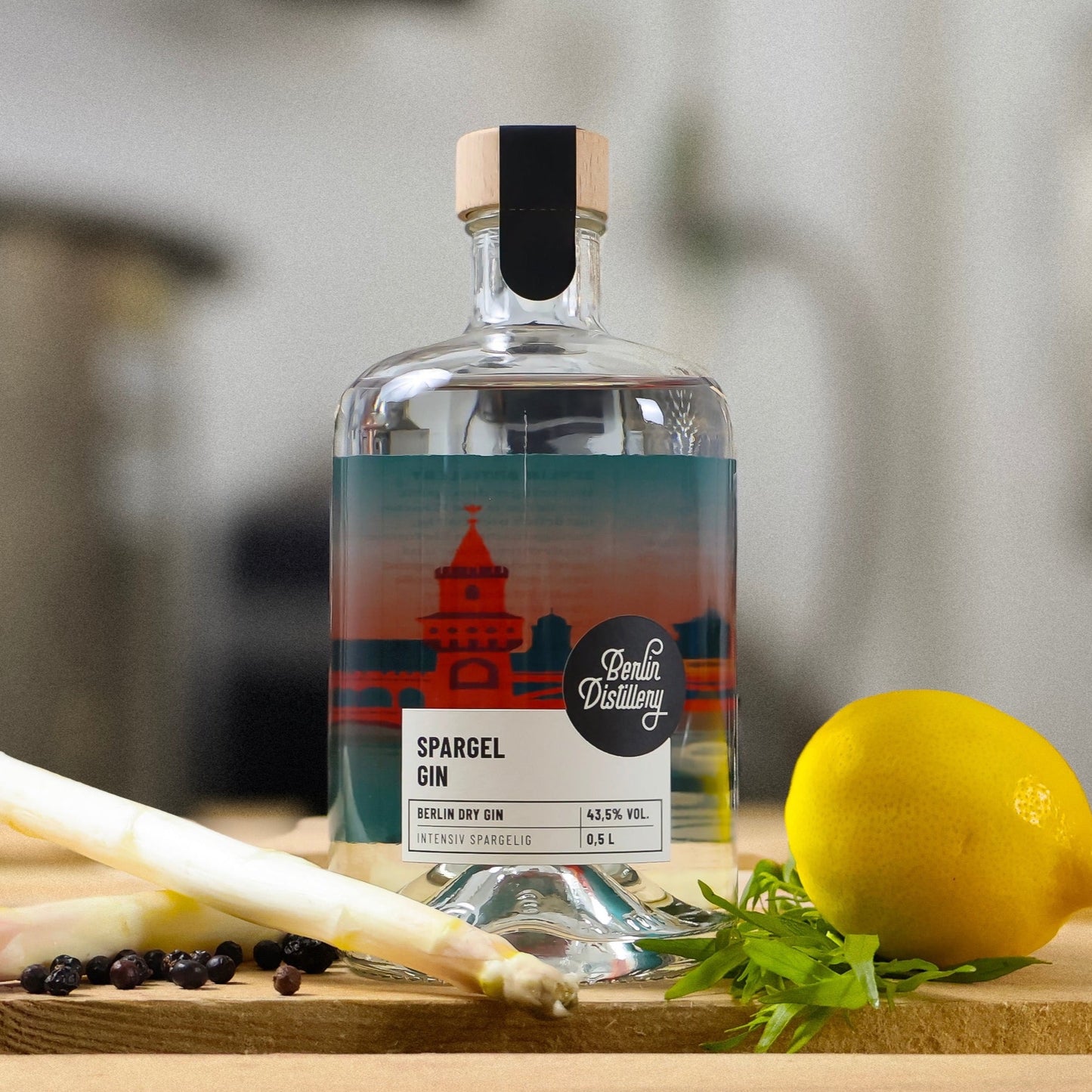 Eine Flasche Berlin Distillery Spargel Gin steht auf einer Holzpalette vor der Produktionsanlage. Neben der Flasche sind weißer Spargel, Wacholderbeeren, eine Zitrone und Estragon zu sehen.