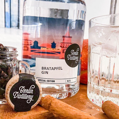 Eine Flasche Berlin Distillery BRATAPFEL GIn steht auf einem Tisch umgeben von Zimtstangen, einem Apfel und einem Glas gebrannte Mandeln.